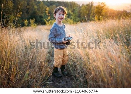Cute toddler boy with old retro vintage camera on autumn grass background. Child with curly hair and grey coat playing with photo