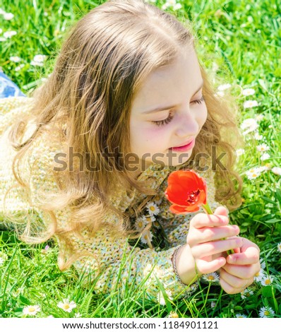 Allergy concept. Girl on happy face holds red tulip flower on sunny spring day. Girl with long hair lying on grassplot, grass background. Child enjoy fragrance of tulip while lying at meadow.
