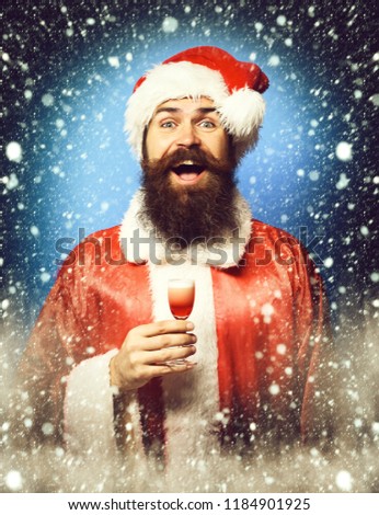 handsome bearded santa claus man with long beard on smiling face holding glass of alcoholic shot in red christmas or xmas sweater and new year hat on blue studio background.