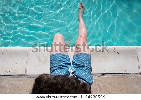 Looking down at man sitting on the edge of the pool with one foot in the water wearing blue swimming trunks. Fit man sits poolside. Caucasian man by an outdoor swimming pool in the summer. Royalty-Free Stock Photo #1184930596