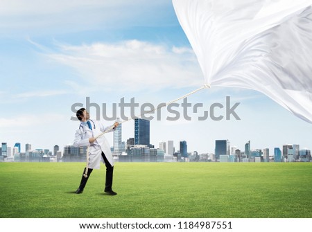 Horizontal shot of young doctor in white medical uniform pulling white fabric while standing on green meadow with cityscape view on background