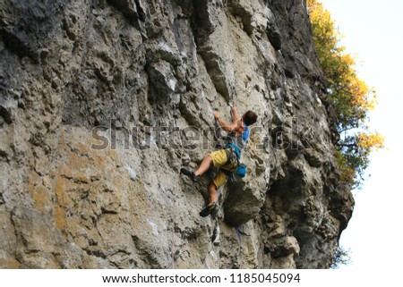 Sports images of a lean muscled rock climber on a broad limestone cliff.  He is sport climbing with safety gear: a rope, quick draws, harness and chalk bag.  It is a bright sunny summer day.