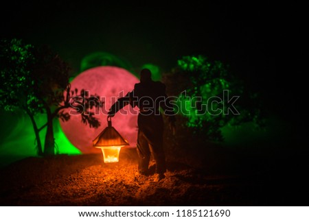 Man in raincoat coming from dark forest with glowing lantern in his hand concept. Silhouette of a horror killer with lamp. Decoration