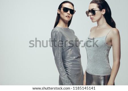 Two women wearing silver trendy outfit with sunglasses standing on grey background. Female model in futuristic look in studio.