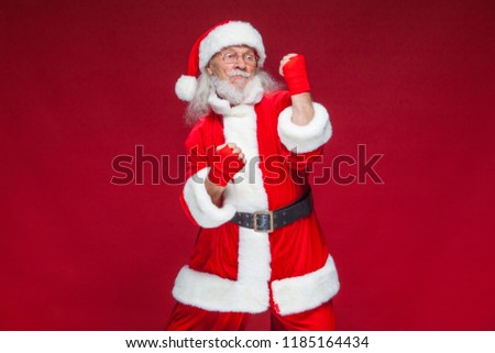 Christmas. Santa Claus with red bandages wound on his hands for boxing imitates kicks. Kickboxing, karate, boxing. Isolated on red background.