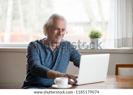 Smiling senior middle aged man in glasses working on laptop at home, happy elderly mature male user looking at computer screen communicating online or using software for dating, reading morning news Royalty-Free Stock Photo #1185179074