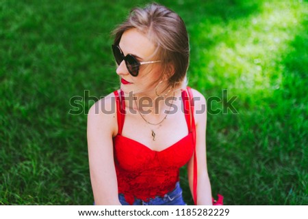 Beautiful happy girl in red tops, blue jeans and sunglasses sits in a park on the grass. Outdoor portrait of young attractive woman.