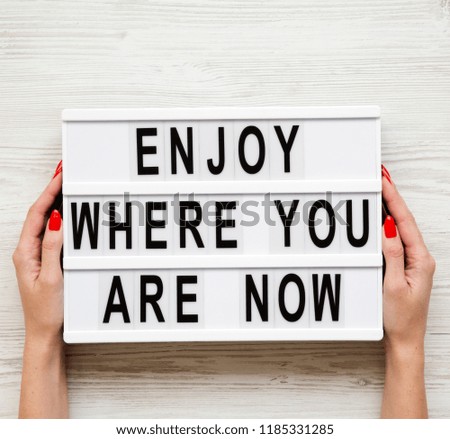 Female hands hold modern board with 'Enjoy where you are now' word over white wooden background, top view. Business concept. From above, flat-lay, overhead. 