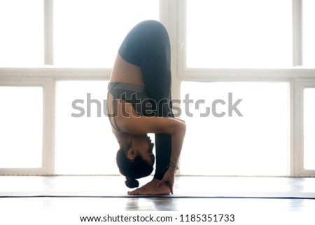 Young sporty attractive woman practicing yoga, doing head to knees exercise, uttanasana, Standing forward bend pose, working out, wearing sportswear, indoor full length, at yoga studio, side view