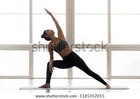 Young sporty attractive woman practicing yoga, doing Reverse Warrior exercise, Viparita Virabhadrasana pose, working out, wearing sportswear, grey pants, top, indoor full length, at yoga studio