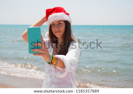 Christmas young smiling woman in red santa hat taking picture self portrait on smartphone at beach over sea background