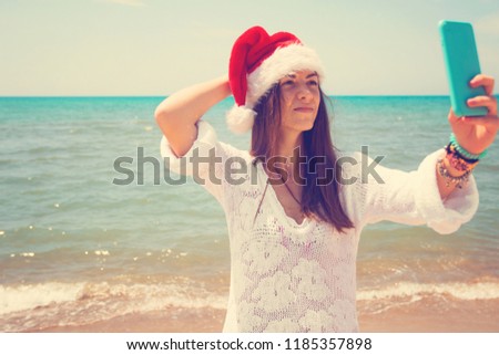 Christmas young smiling woman in red santa hat taking picture self portrait on smartphone at beach over sea background