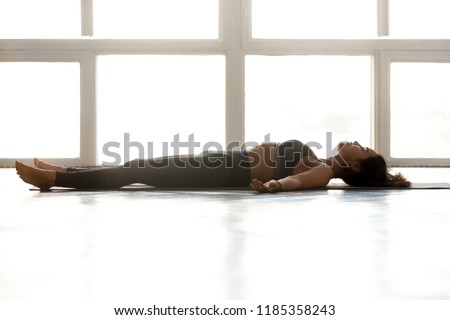 Young sporty attractive woman practicing yoga, doing Dead Body exercise, Savasana, Corpse pose, working out, wearing sportswear, grey pants, top, indoor full length, at yoga studio, side view Royalty-Free Stock Photo #1185358243