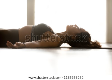 Young sporty attractive woman practicing yoga, doing Dead Body exercise, Savasana, Corpse pose, working out, wearing sportswear, grey top, indoor close up, at yoga studio, side view Royalty-Free Stock Photo #1185358252