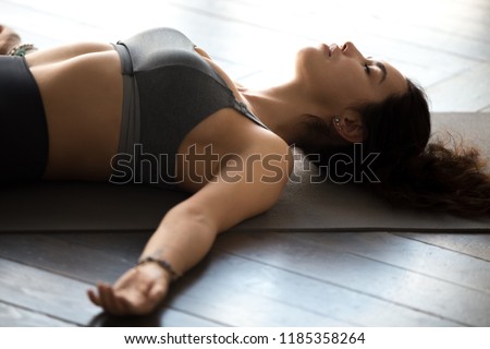 Young sporty attractive woman practicing yoga, doing Dead Body exercise, Savasana, Corpse pose, working out, wearing sportswear, grey top, indoor close up, at yoga studio Royalty-Free Stock Photo #1185358264