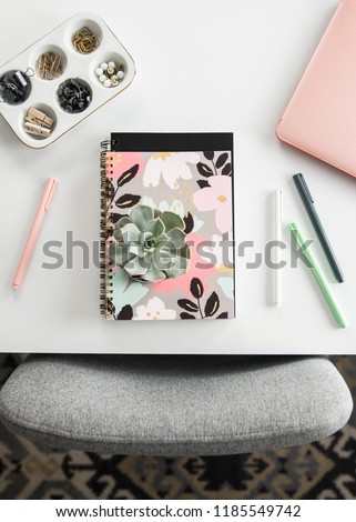 A birds-eye view of a feminine styled office with a white desk, rose gold laptop, flower notebook, and a succulent.