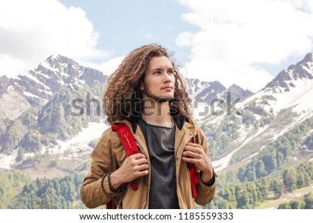 Young curly haired Man Traveler with red backpack relaxing outdoor with rocky mountains on background
