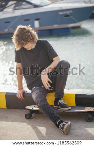 handsome man sitting with longboard on street on pier with yacht