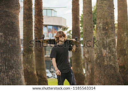Handsome skateboarder wering sunglasses holds longboard on shoulder on city street or tropic background