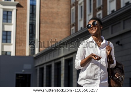 Happy relaxed young African female student in white basic shirt and trendy sunglasses with leather backpack looking at her smartphone in summer urban street over modern business centre buildings.