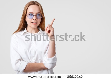 Waist up shot of pretty woman in blue shades, shows direction with index finger, dressed in casual white t shirt, isolated over studio background, Free space for your promotion, advertisement