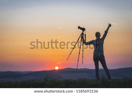 Female photographer holding a tripod with camera and looking at sunset. Woman enjoying spending time with camera in nature.