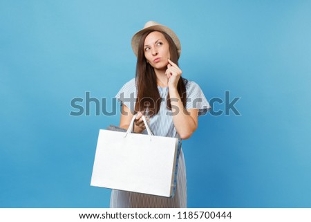 Portrait pensive beautiful caucasian woman in summer dress, straw hat looking up, holding packages bags with purchases after shopping isolated on blue pastel background. Copy space for advertisement