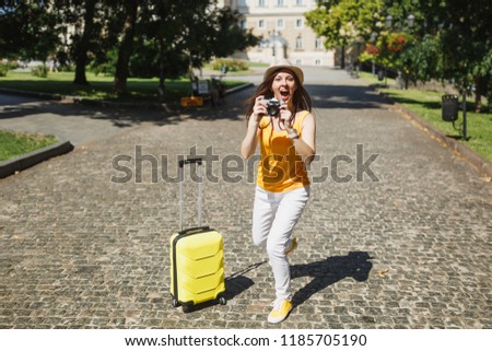 Excited traveler tourist woman in yellow casual clothes with suitcase take pictures on retro vintage photo camera running outdoor. Girl traveling abroad on weekend getaway. Tourism journey lifestyle