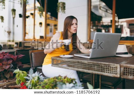 Upset sad girl in outdoors street cafe sitting with laptop pc computer, looking on mobile phone texting message sms, disturb of problem, in restaurant during free time. Mobile office freelance concept