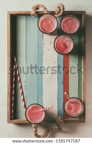Red fruit berries watermelon iced cocktail in glasses with striped straw on vintage wooden tray over white marble background. Summer cold drink. Flat lay, space