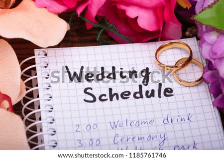 Two Golden Rings With Wedding Schedule On Spiral Notepad Surrounded By Flowers