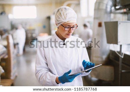 Young focused female worker in sterile clothes checking productivity of production line in food factory.