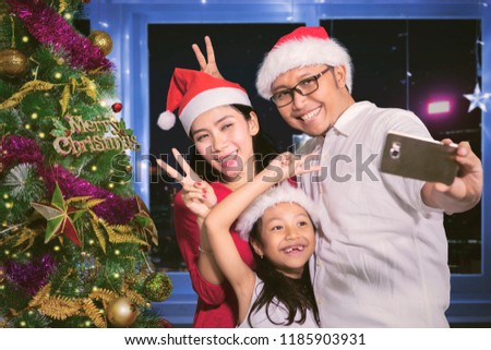 Portrait of young parents with their daughter taking a selfie photo together by using a smartphone near a Christmas tree in the living room