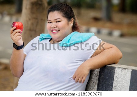 Picture of happy obese woman holding a fresh apple while sitting in the park