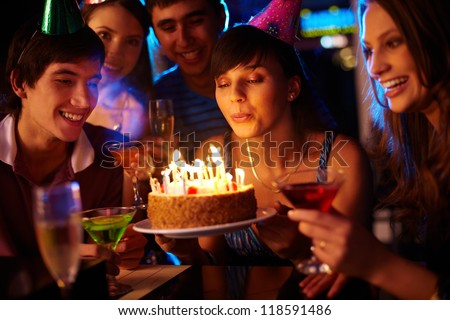 Portrait of charming girl blowing on candles on birthday cake surrounded by friends at party