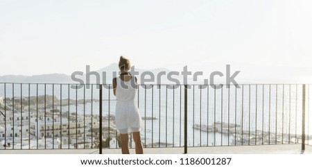 A young girl takes pictures of a Greek island from the balcony. White houses on the shore, yachts in the sea