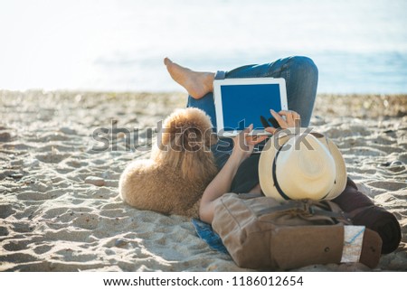 Woman traveler is holding touch pad, while is relaxing outdoors during his trip on the sea. Woman is traveling with her dog.