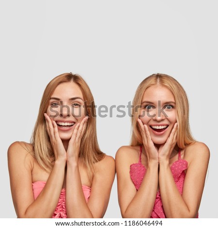 Vertical portrait of joyful two girls with broad smiles, being amazed as see something incredible and nice, stand next to each other, isolated over white background. Positive emotions concept