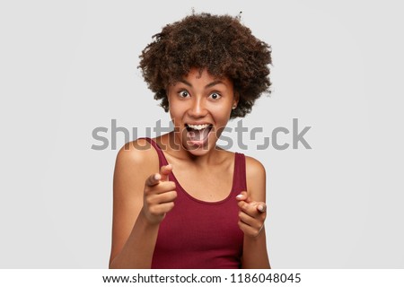 Cheerful attractive dark skinned young girlfriend points to you, has joyful look at camera, poses against white background, dressed in casual t shirt. Ethnicity, body language and choice concept