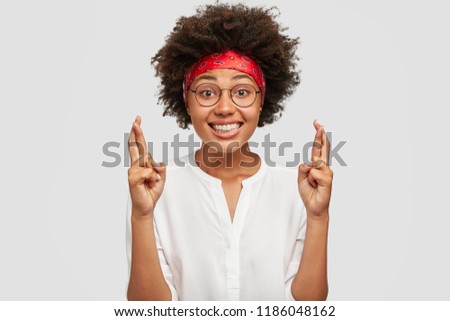 Shot of cheerful carefree dark skinned woman keeps hands raised, fingers crossed, makes wish, dreams about something, has curly hair, broad smile, isolated over white background. Body language concept