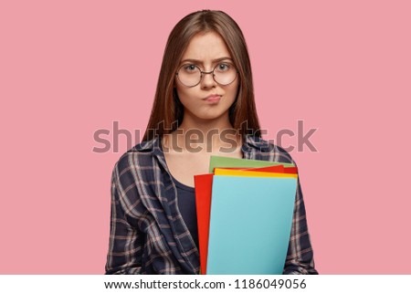 Clueless female reader purses lips with doubtful expression, has dark straight hair, wears transparent glasses, has gloomy expression, poses against pink studio wall. Education and facial expressions