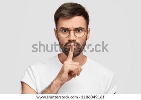Shush, calm down please! Serious strict young teacher demands keep voice down, shows silence gesture, dressed in white t shirt, conducts lesson for pupils, stands indoor. Man tells secret to friend