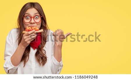 Lunch time concept. Attractive cheerful woman eats delicious Italian pizza, surprised with wonderful taste, indicates with thumb aside, shows where pizzeria situated, suggests to visit and have snack Royalty-Free Stock Photo #1186049248