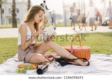 Pretty young girl having a picnic in a public parc, reading a book and enjoying good weather after taking pictures on her camera. Sunny weather, basket with food for picnic, summer holidays concept 