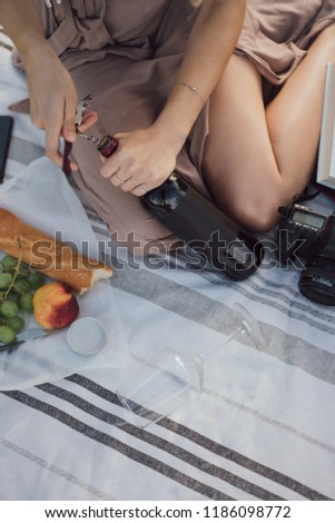 Top view cropped picture of a girl opening a bottle of blank red wine while having a picnic in a city park sitting on a green grass and a cotton cover with fruit above. Mockup of a wine bottle