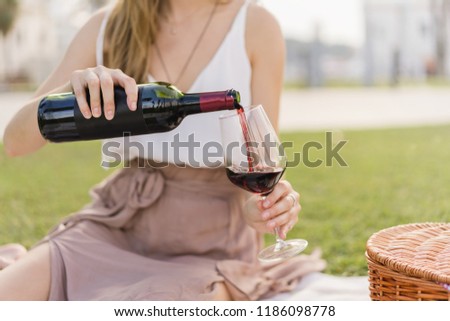 Cropped picture of a girl pouring wine sitting on a green grass outdoors. Concept of having picnic in a city park during summer holidays or weekends. Blank bottle of wine with a space for your logo