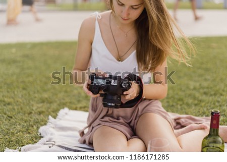 Young girl taking photos on a dslr camera while having a picnic in the park with wine, looking on the screen at pictures, female photographer, green grass on the background, shooting outdoors