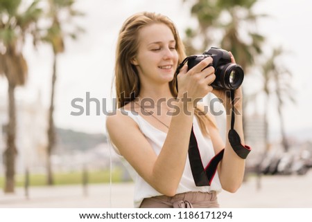 Pretty young girl laughing while taking pictures on professional dslr camera walking outdoors in the park on the summer sunny day, traveling, making photos, photography as a hobby, looking into camera