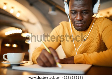 Young serious man with headphones looking at blank paper in front of him while learning to draw professionally