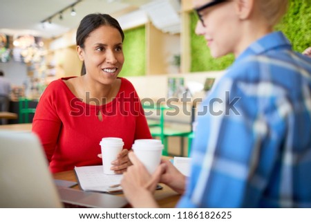 Young female employee in red casualwear looking at her colleague during discussion of online financial rates in cafe